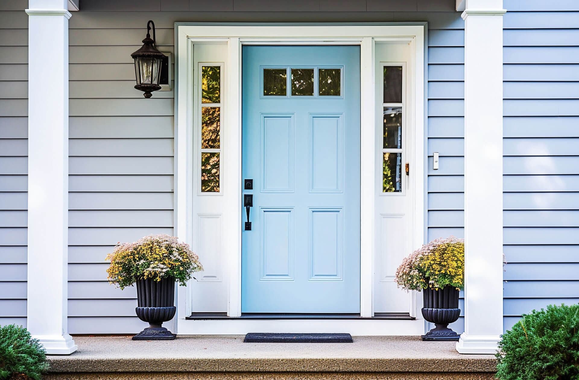 Light blue front entry door with glass sidelights, white trim, gray siding, porch columns, and matching flower planters.
