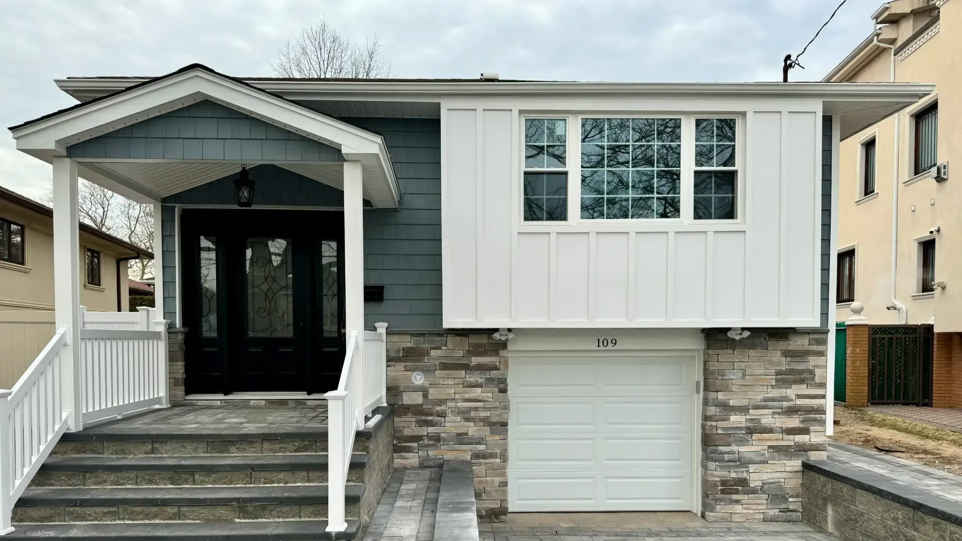 Renovated home exterior with blue siding, white board-and-batten accents, stone veneer, new windows, covered entry, and garage.