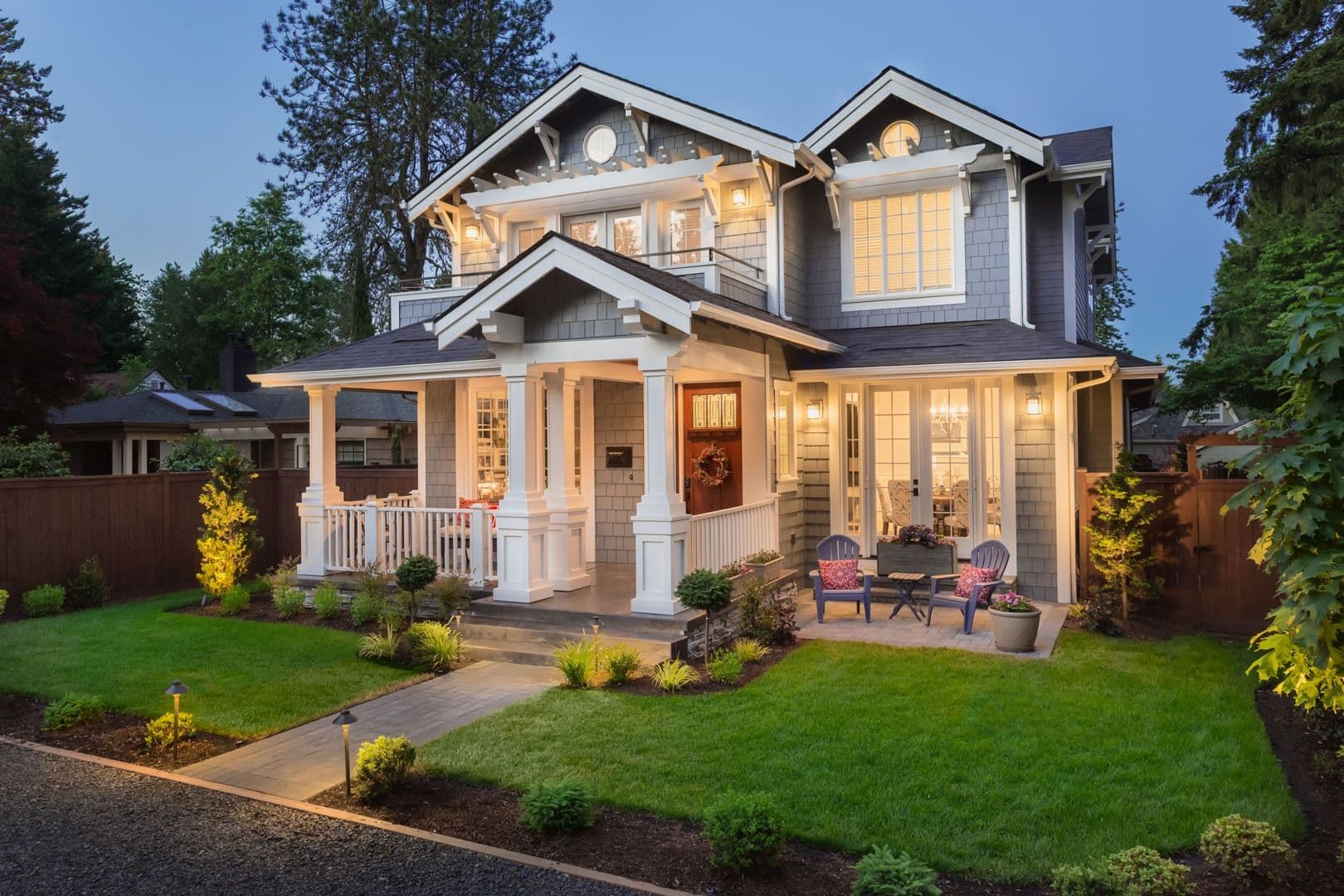 Two-story gray Craftsman-style home with white trim, illuminated windows, a covered front porch, and landscaped lawn at dusk.