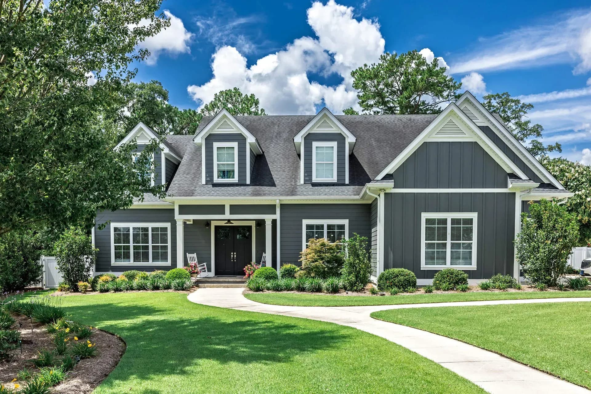 Large gray Craftsman-style home with white trim, dormer windows, a covered front porch, and a landscaped lawn.