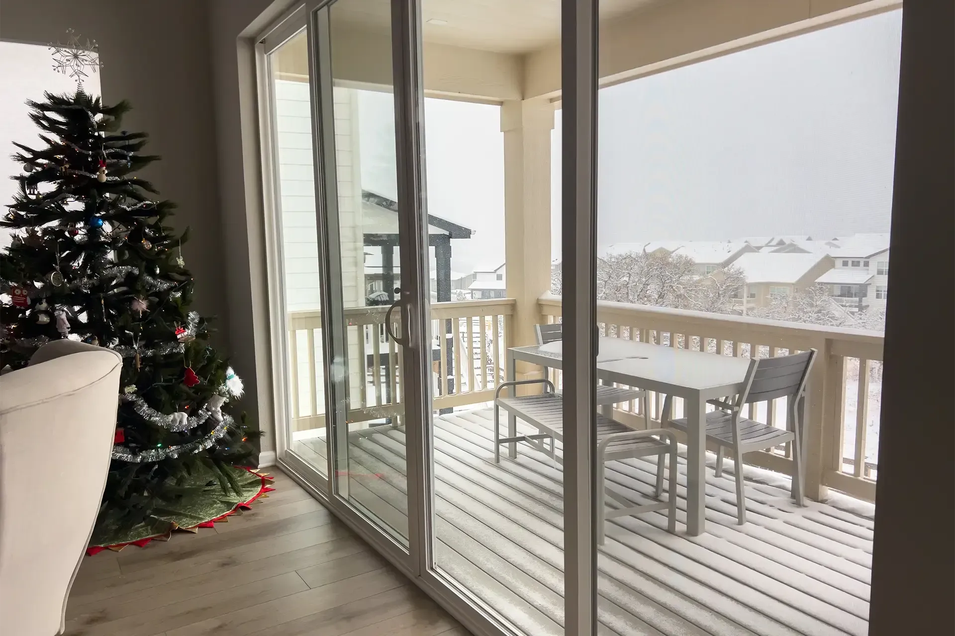 Sliding glass patio doors overlooking a snow-covered balcony with outdoor furniture and a decorated Christmas tree indoors.