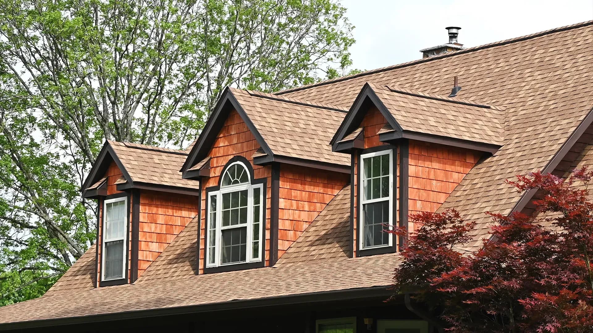 Home exterior with cedar shake siding, brown asphalt shingle roof, and three dormer windows with white trim.