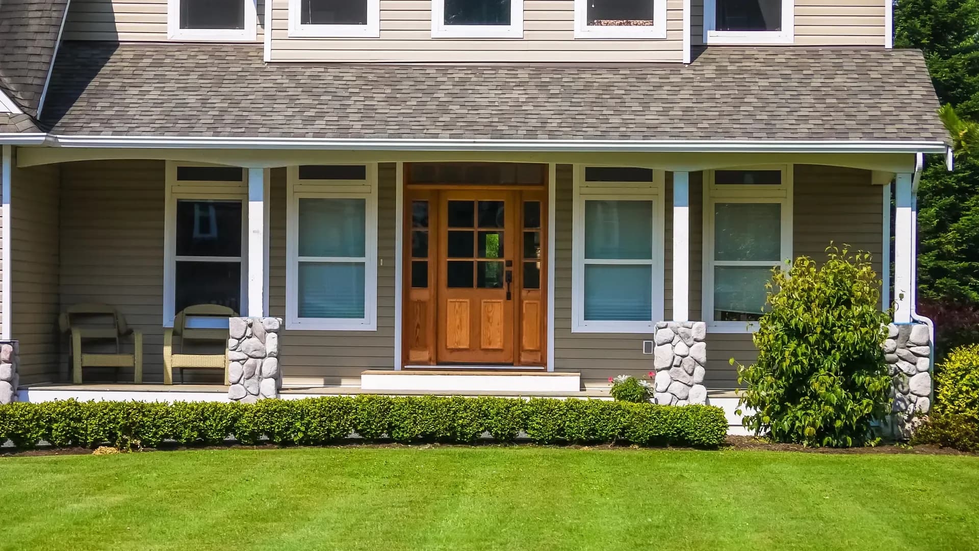 Front exterior of a home with tan siding, white-trimmed windows, a wooden entry door, stone porch columns, and landscaped lawn.