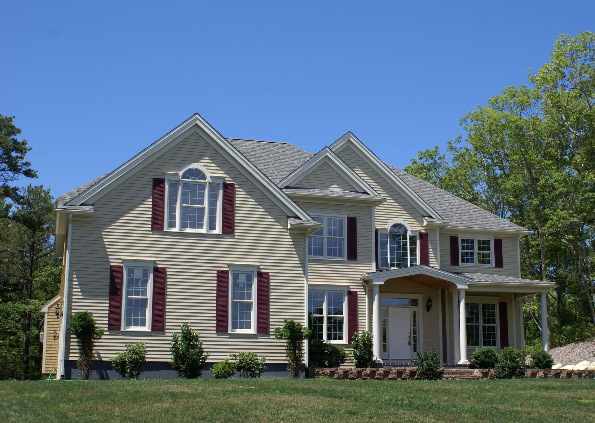 Two-story home with beige siding, burgundy shutters, white-trimmed windows, a covered front entry, and a landscaped lawn.