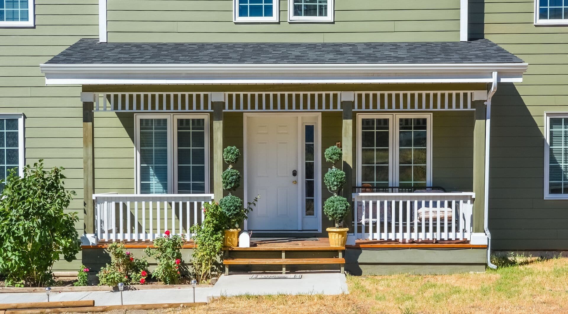 Front porch of a green-sided home with white railings, a white entry door, potted topiary plants, and flower beds.