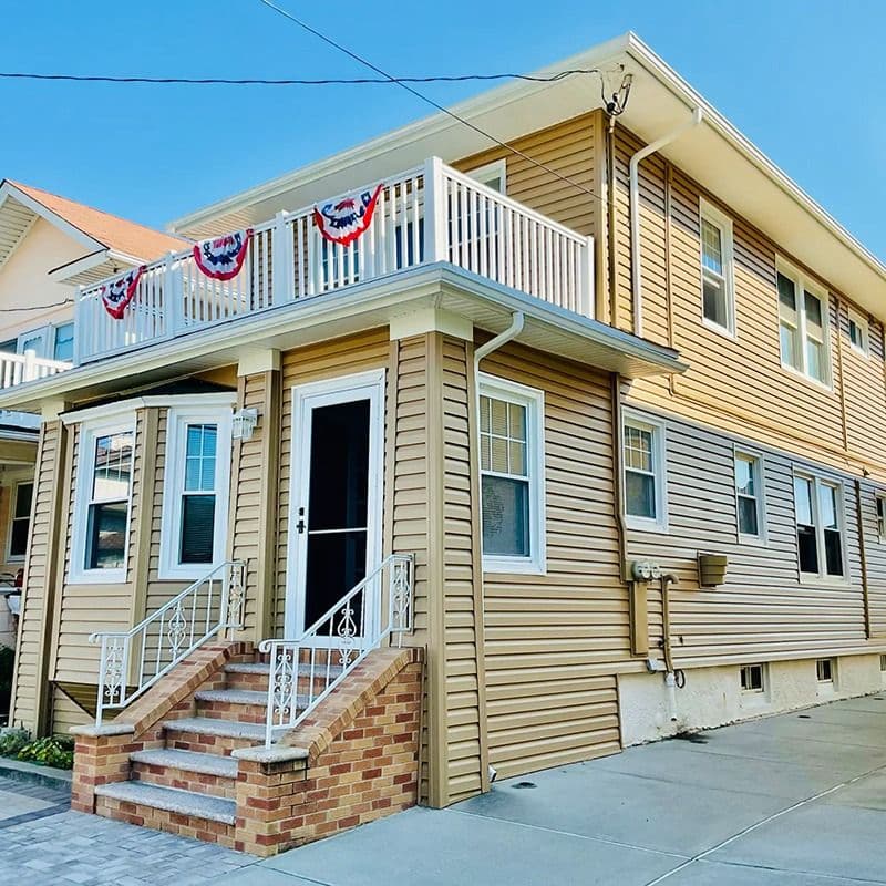Two-story home with tan vinyl siding, white-trimmed windows, brick front steps, and an upper-level balcony.