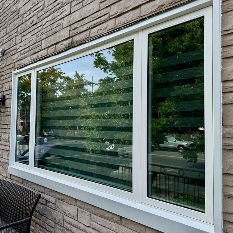 Large white-framed vinyl picture window installed in a stone exterior wall, reflecting trees and the street.
