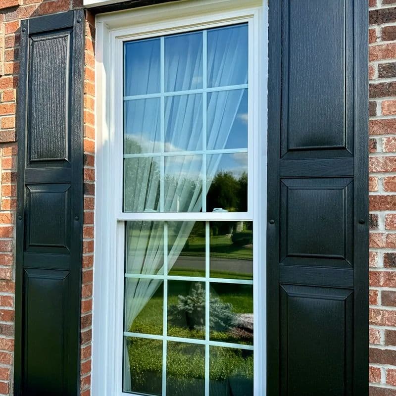 White-framed double-hung window with divided panes and black shutters installed in a red brick wall.