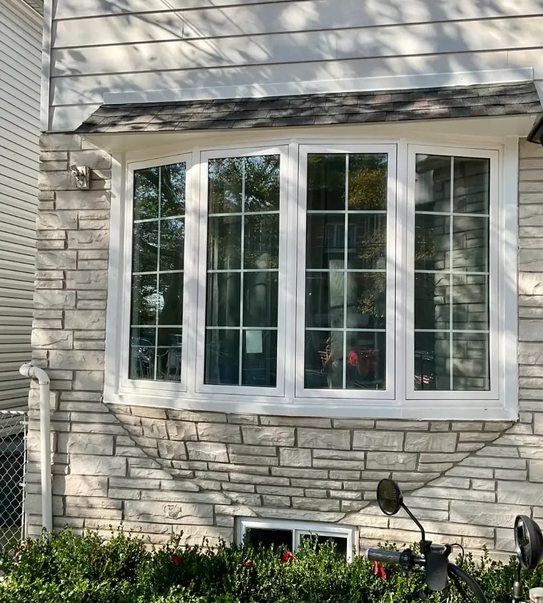 White bow window with divided panes installed in a light stone exterior wall beneath a small shingle awning.