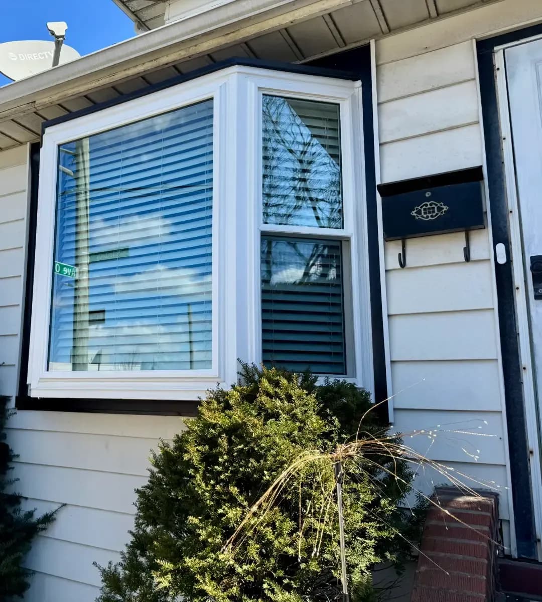 White-framed bay window with blinds installed on a light-colored home exterior beside a black mailbox.