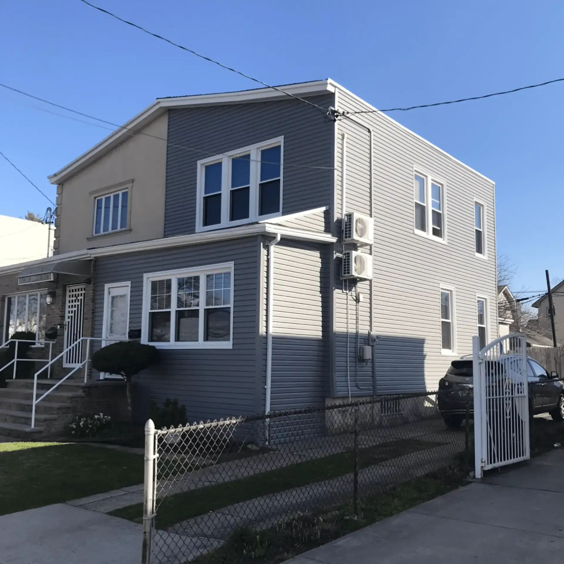 Exterior view of a large two-story house featuring contrasting dark and light grey horizontal lap siding, white window trim, a chain-link fence, and a white driveway gate.