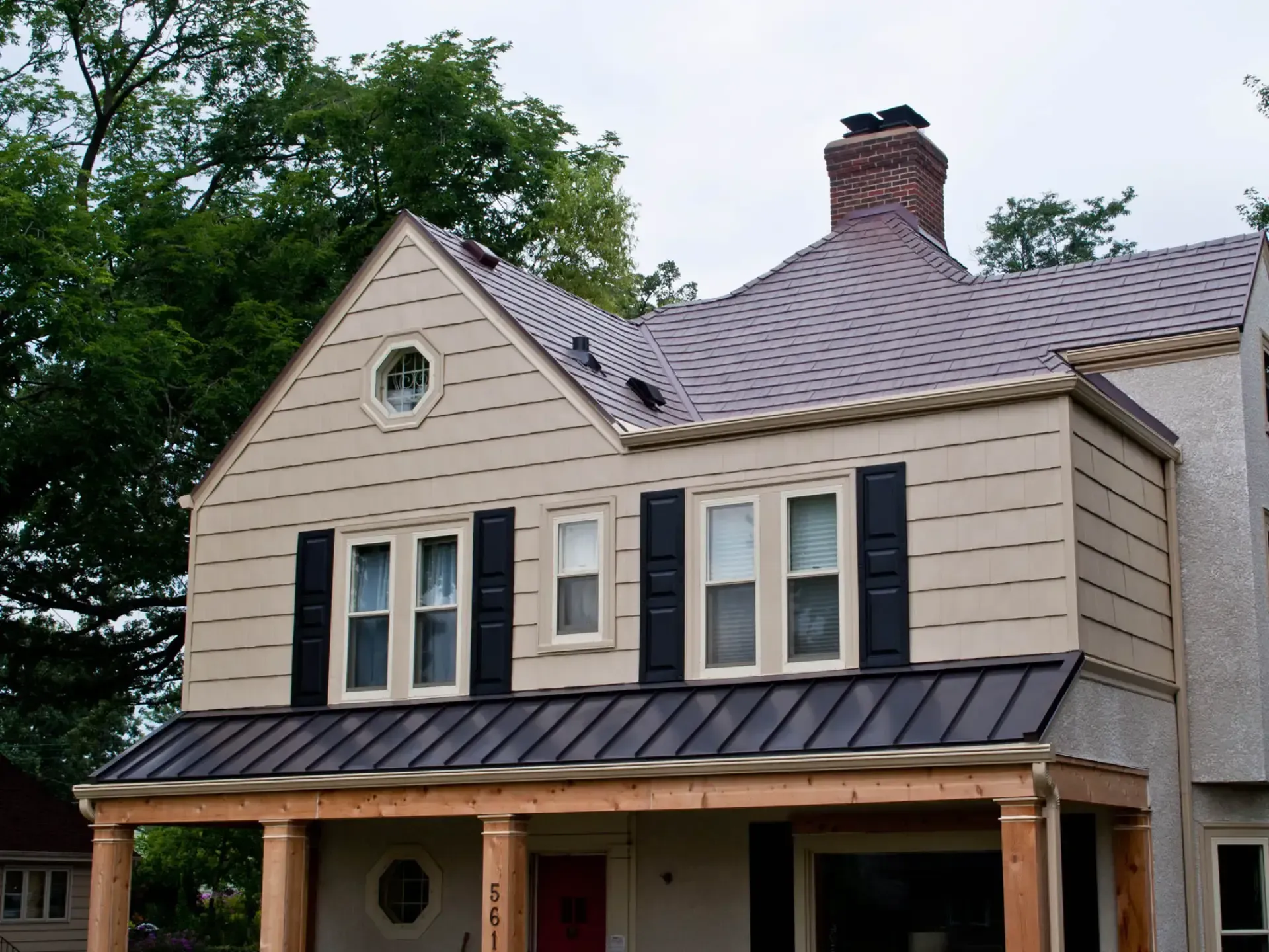 Exterior view of a two-story beige-sided house featuring a dark brown slate-style main roof, black shutters, an octagonal window, and a dark brown standing-seam metal awning over a timber front porch.
