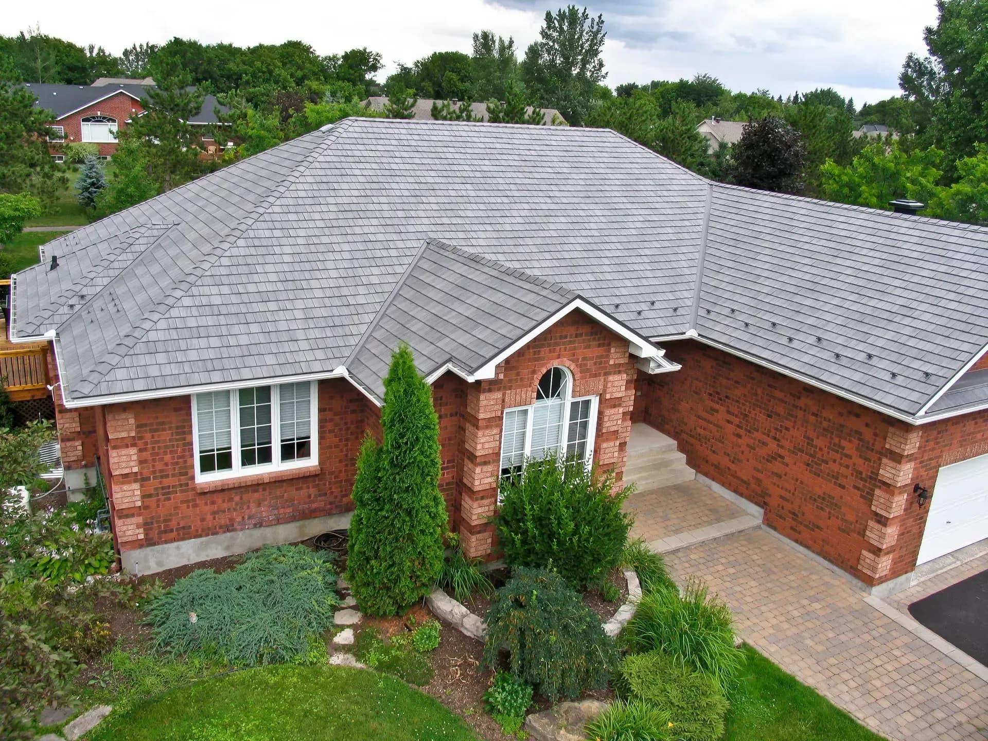 High-angle aerial view of a single-story red brick home featuring a multi-gabled light grey slate-style shingle roof, a paved brick driveway, and manicured evergreen landscaping.