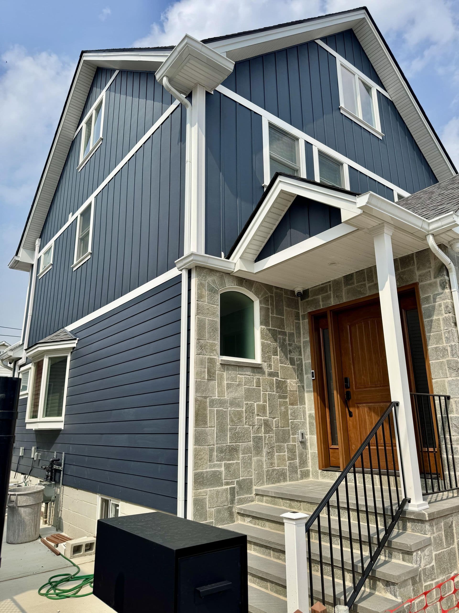 Low-angle exterior view of a multi-story home featuring dark navy blue vertical board-and-batten siding on the upper floors, horizontal lap siding on the lower level, crisp white trim, and a grey stone masonry entry porch with a warm wood front door.
