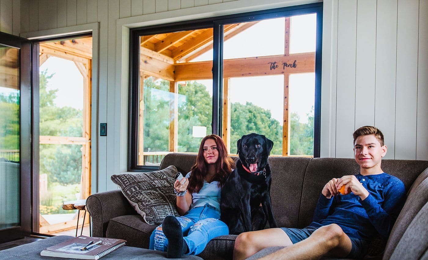 Interior view of a cozy living room featuring a large dark-framed sliding window overlooking a wooden porch structure. A young woman holding a remote, a black dog, and a young man holding an orange are sitting together on a dark grey sofa in front of the window.