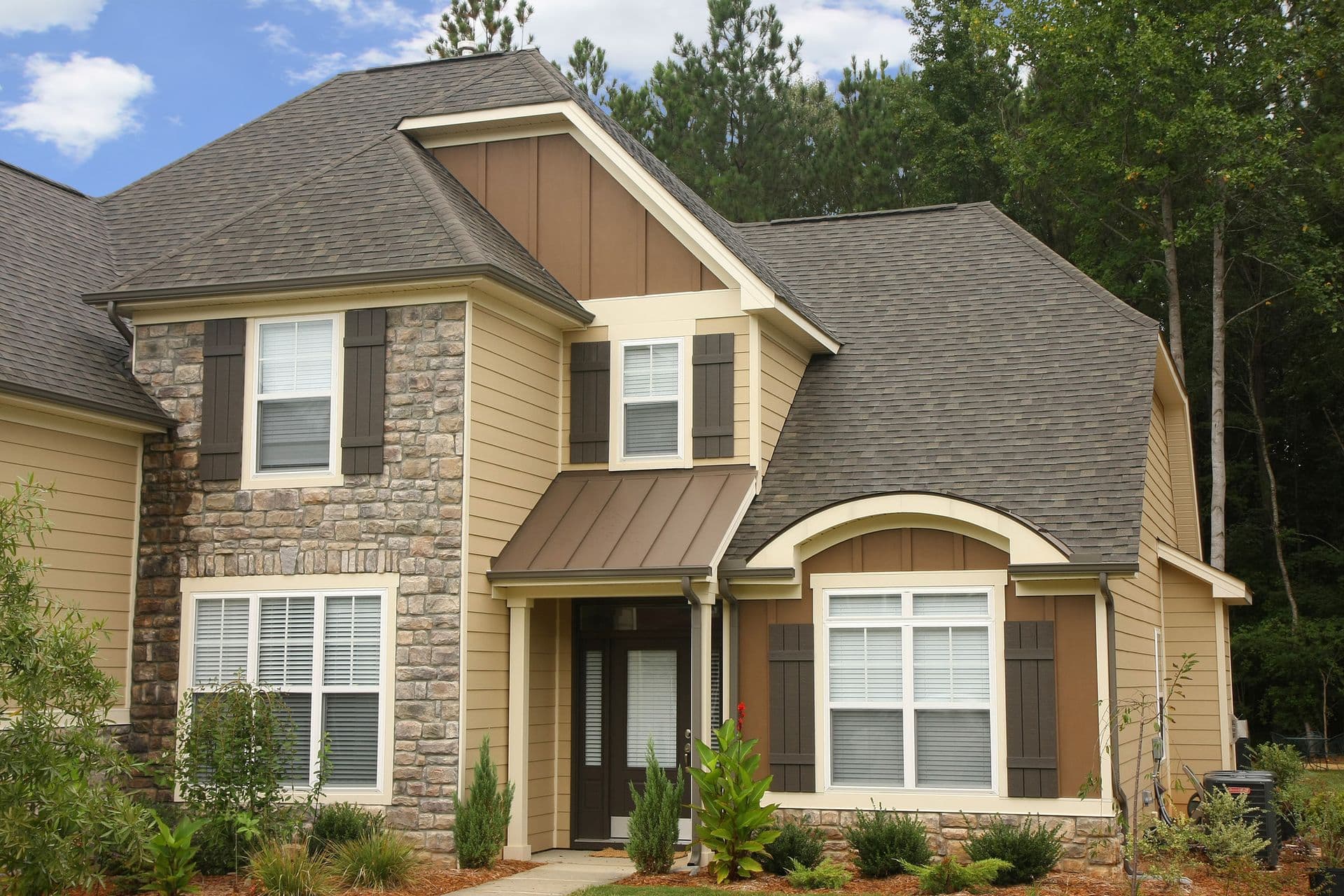 A bright, high-angle exterior view of a multi-gabled, two-story home featuring light tan horizontal lap siding, brown board-and-batten accent panels in the gables, rustic stone veneer sections, dark brown raised-panel window shutters, a brown standing seam metal awning over the front entrance, and weathered wood asphalt roof shingles.