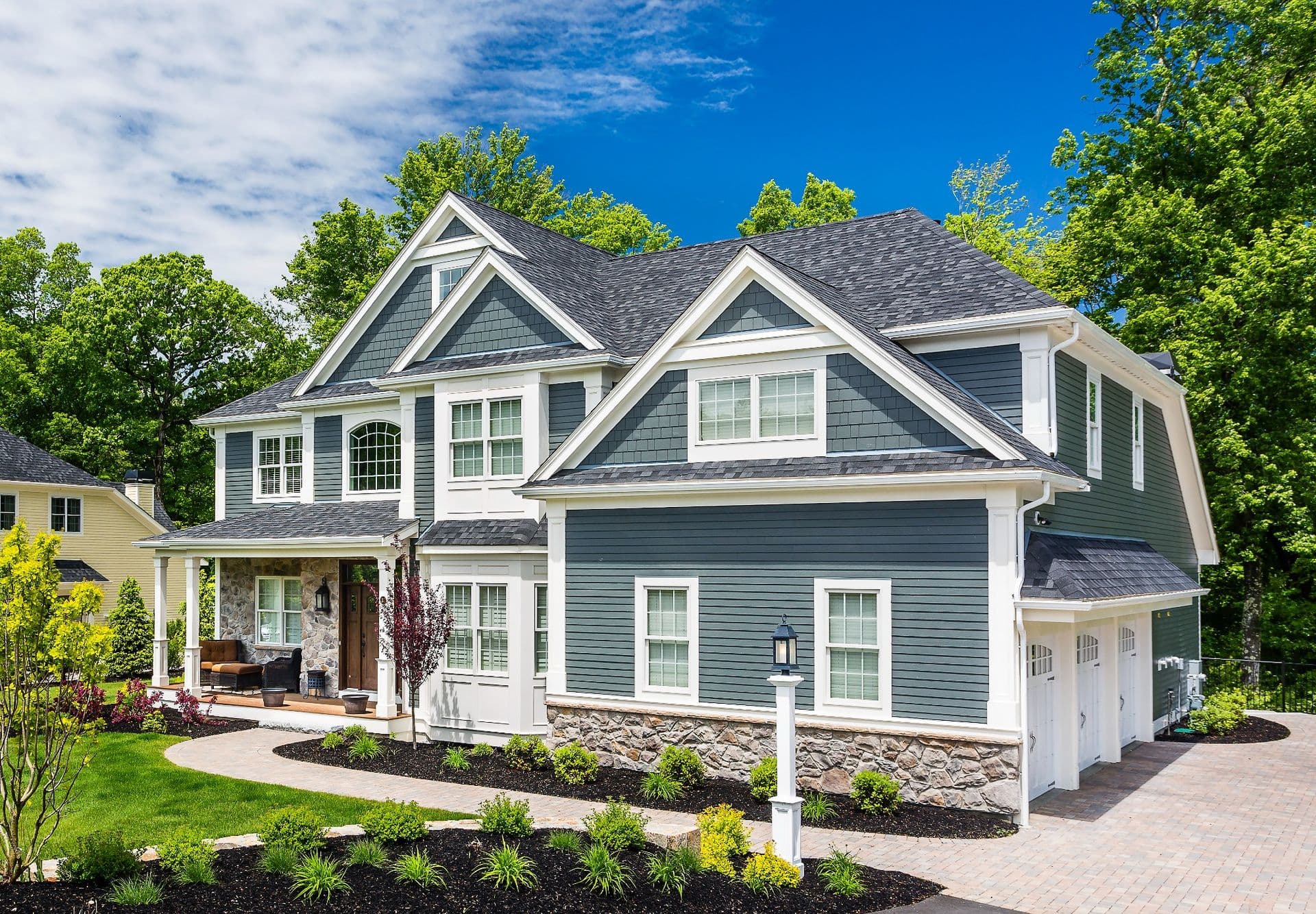 Large Craftsman Home with Slate Blue James Hardie Siding (Most-Asked-Questions-About-James-Hardie-Siding.jpg) A bright exterior view of a large two-story Craftsman-style home featuring slate blue James Hardie lap and shingle siding, white trim, stone veneer accents along the foundation and front porch, a dark shingled roof, a stone walkway, and a multi-car garage on a sunny day.