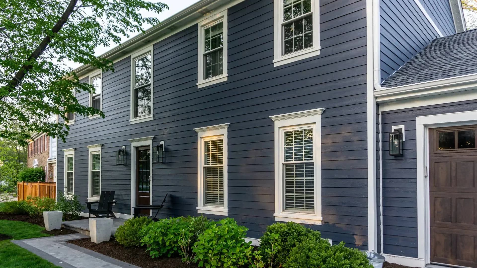 Exterior view of a two-story home clad in dark slate blue James Hardie fiber cement lap siding, accented by crisp white window trim, white gutters, black wall-mounted lanterns, a dark wood front door, and manicured garden shrubbery along a paver walkway.