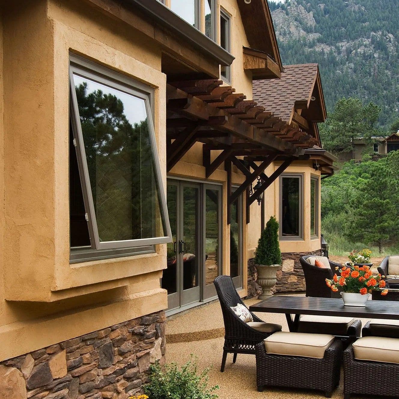 Open dark-framed awning window on a beige stucco house exterior overlooking a stone patio with outdoor wicker dining furniture and a forested mountain background.