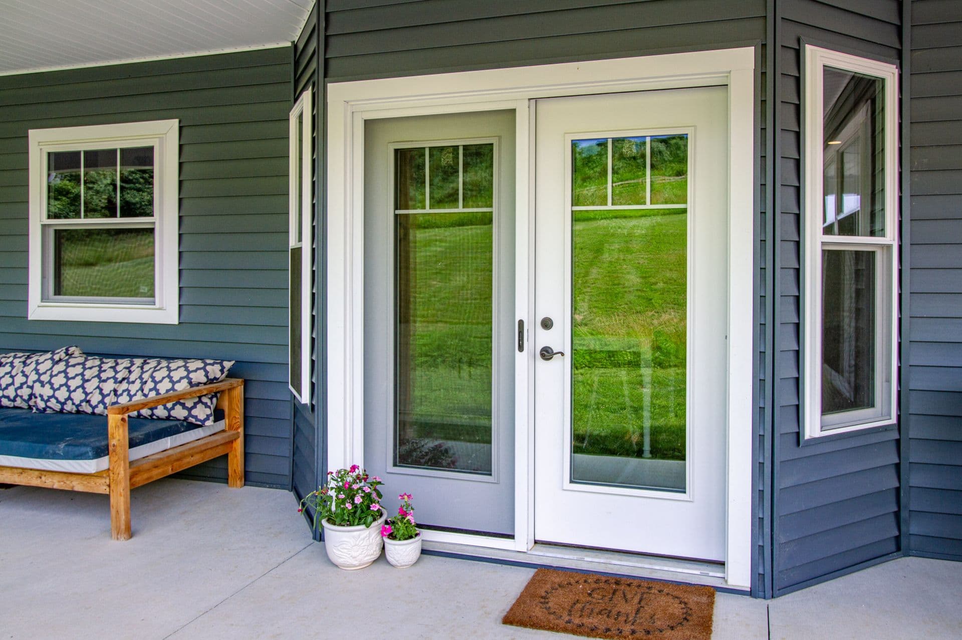 Exterior view of a covered concrete patio surrounded by dark slate-blue horizontal siding. At the center, a white hinged patio door features a large glass panel with upper grilles, paired with a matching sliding screen door. The doorway is framed by thick white molding, with two potted pink flowers and a brown "Give Thanks" doormat at the threshold. To the left, a wooden daybed with blue patterned cushions sits below a white-framed single-hung window, while narrow angled windows flank the entry frame on either side.