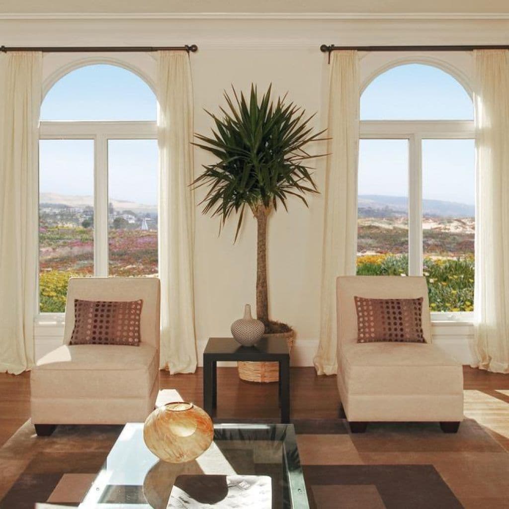 An interior view of a bright, sunlit sitting room featuring two large arched window combinations with white frames and clear glass. The windows offer a scenic view of a sunny landscape.