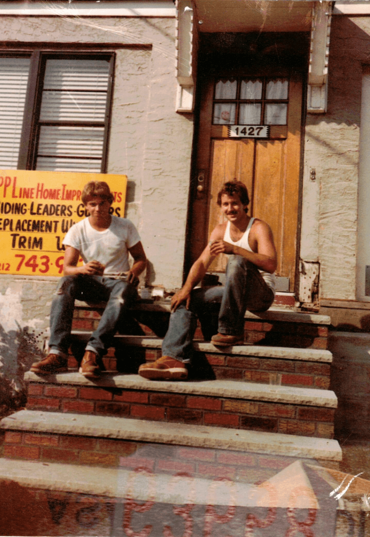 Two workers taking a break on brick steps outside a house with a Top Line Home Improvements sign nearby.