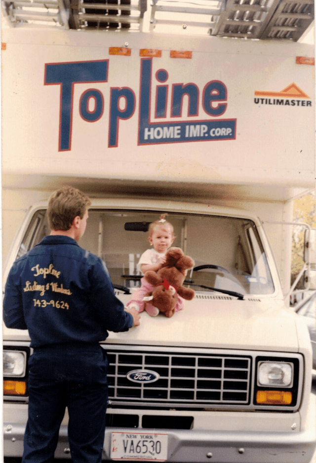 Worker standing next to a TopLine truck with a young child sitting on the hood holding a teddy bear.
