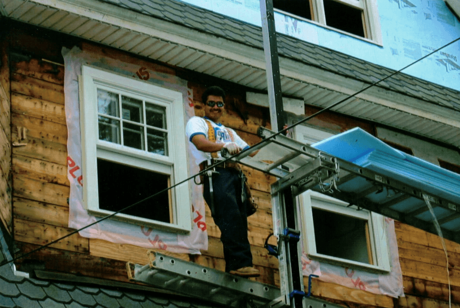 Contractor standing on scaffolding while installing a new double-hung window on a house under renovation.